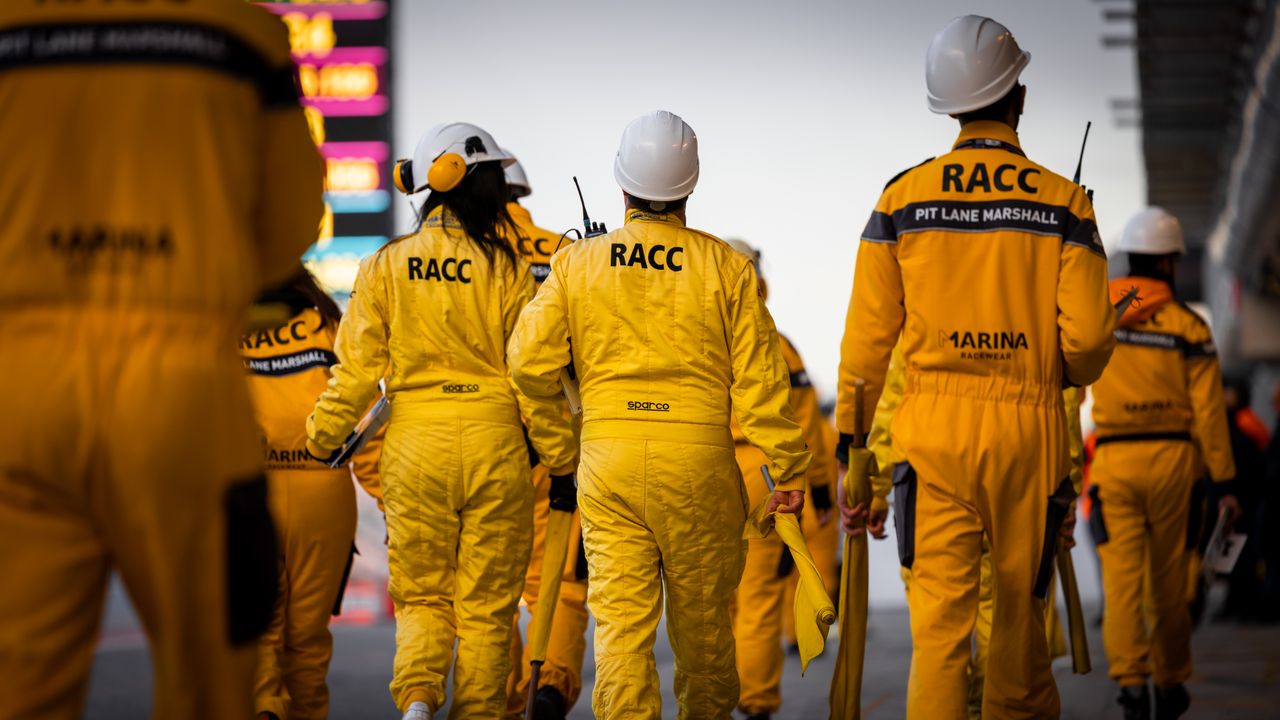 Racing marshals in yellow uniforms walking along the pit lane of a motorsport event.