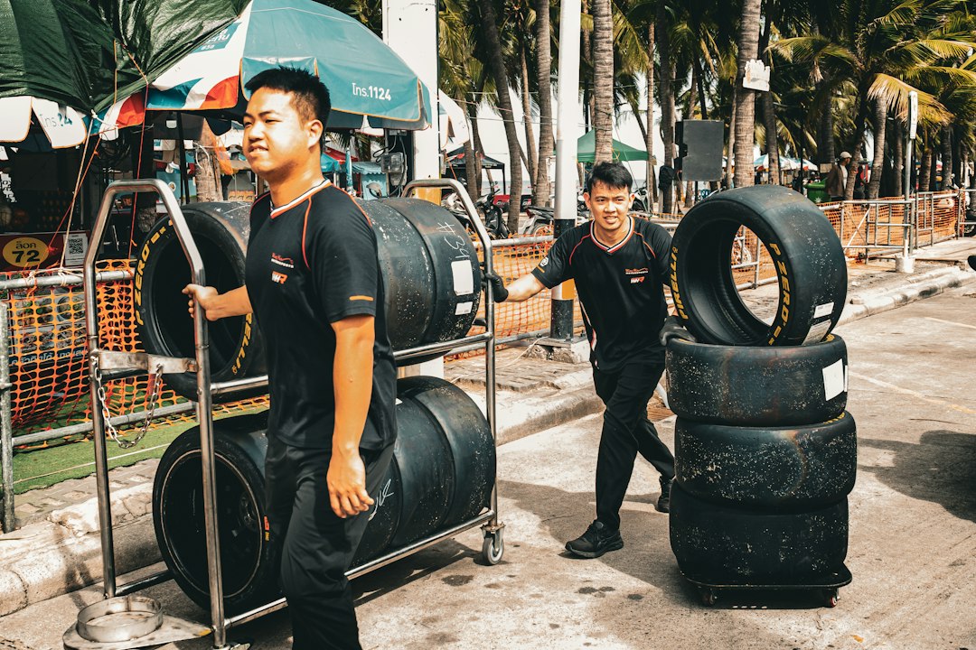 A candid, action-packed photograph of two race team mechanics hauling stacks of Pirelli P Zero racing slick tires on heavy-duty hand carts and dollies through the open-air paddock lane. Set against the tropical, palm tree-lined backdrop of the Bangsaen Street Circuit in Chonburi, Thailand, during the 2026 Bangsaen Grand Prix / Thailand Super Series. This image captures the human effort, tire management strategy, and vibrant coastal atmosphere of Asian motorsport logistics.